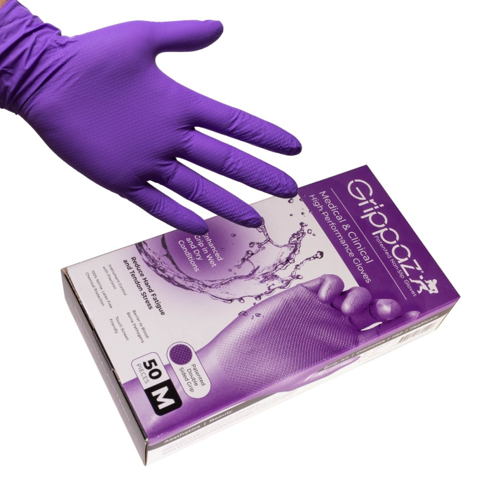 Une main portant un gant jetable violet plane au-dessus d'une boîte de Boite de 50 gants Grippaz, gants antidrapants haute performance en nitrile violets, avec des graphiques d'éclaboussures d'eau et des détails sur le produit visibles sur l'emballage.